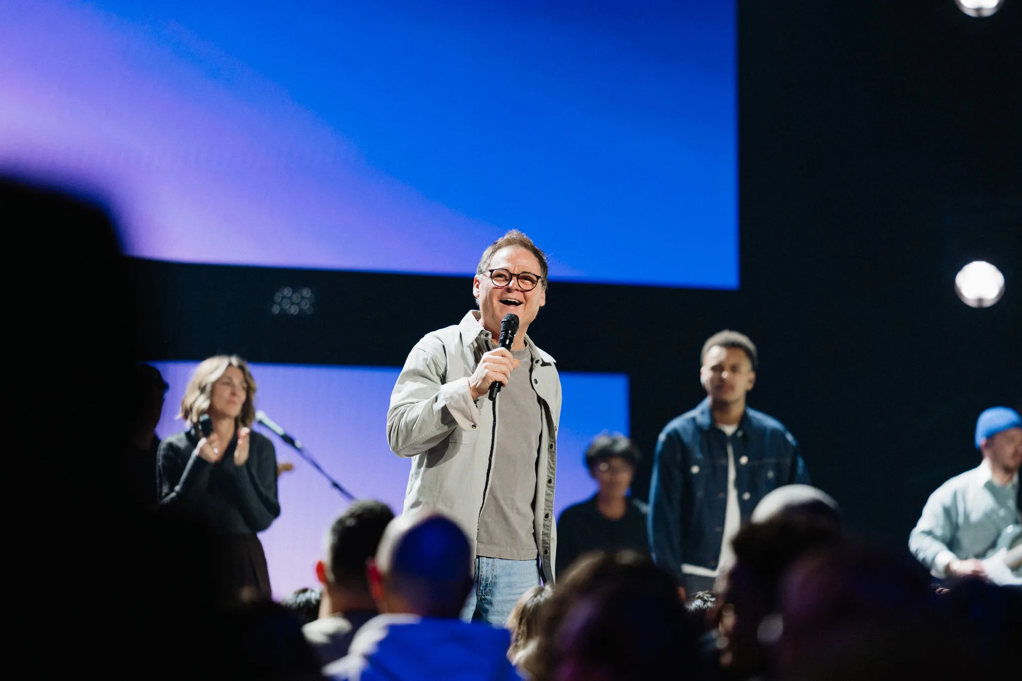 A man speaks into a microphone on stage with people gathered around him under blue stage lighting.