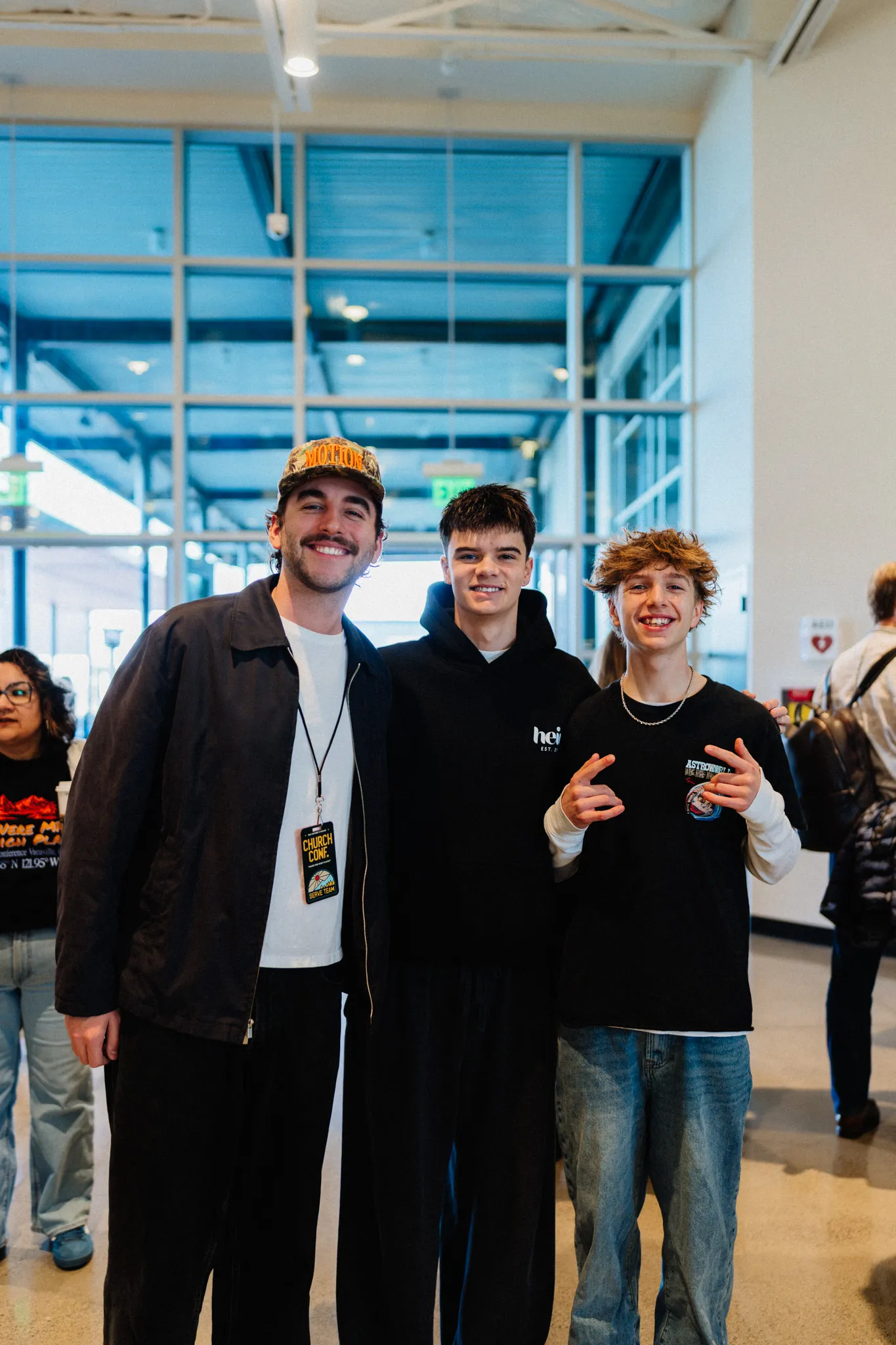 Three young men smile together for a photo in a lobby, one wearing a conference lanyard.
