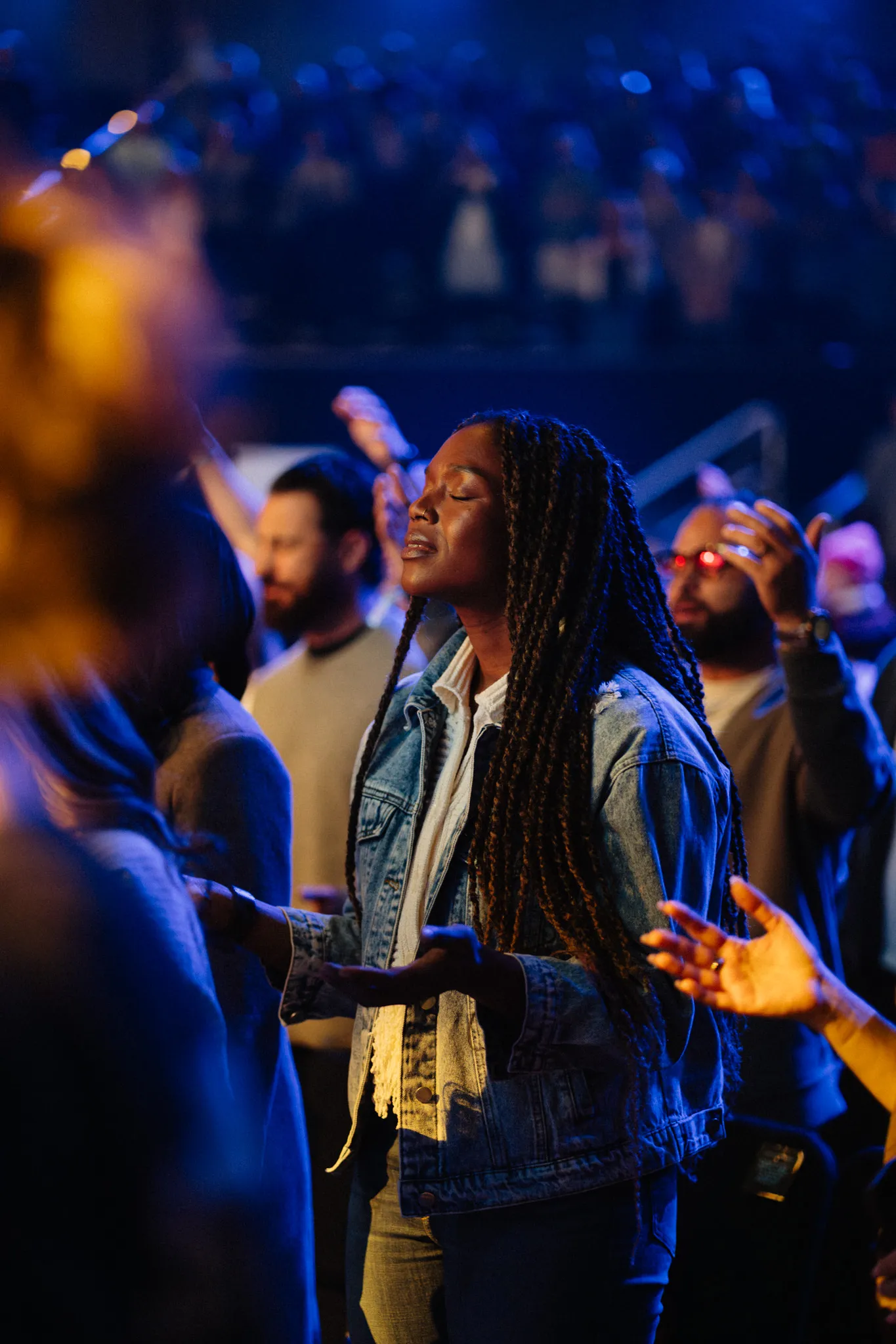 A woman with braided hair worships with her hands raised, eyes closed, in a blue-lit crowd.