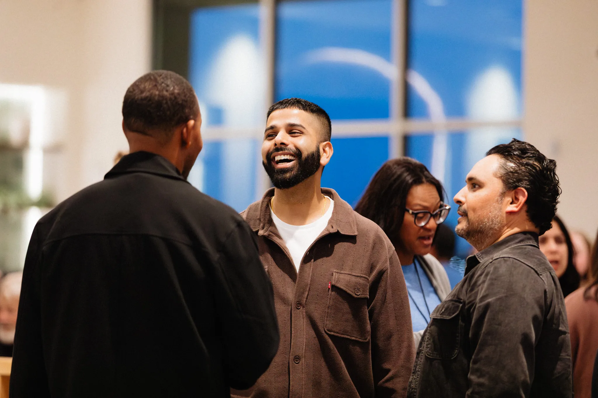A group of people talk and laugh together in a lobby with large windows.