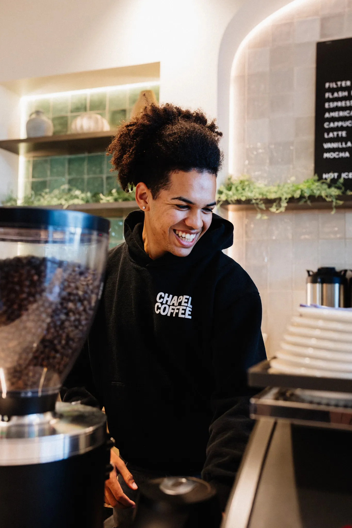 A barista smiles while making a drink at a coffee counter, wearing a "Chapel Coffee" hoodie.