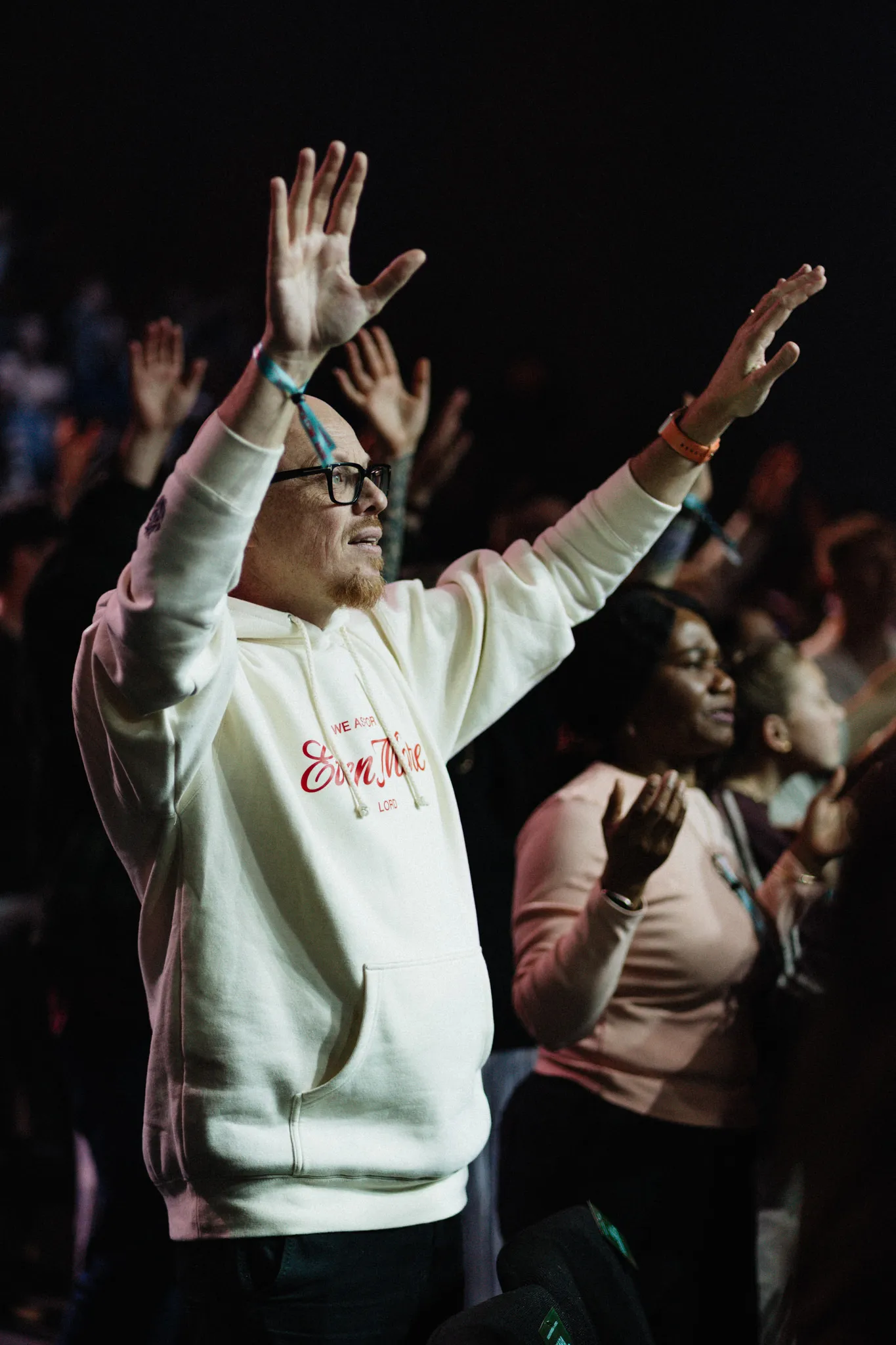 A man in a cream hoodie raises both hands in a worshipping crowd.