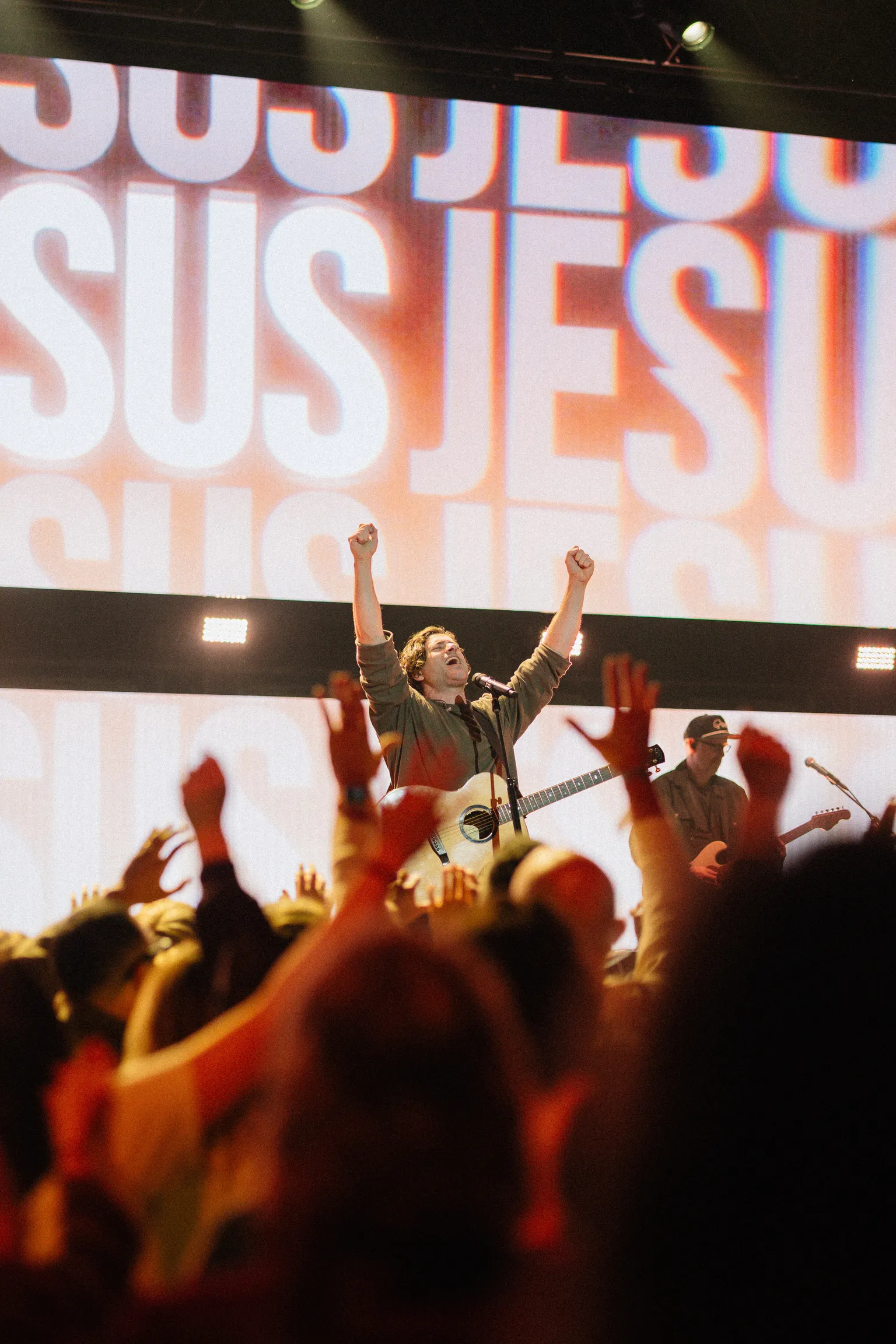A man on stage raises both fists in celebration in front of a screen reading "JESUS," with a crowd raising their hands in the foreground.