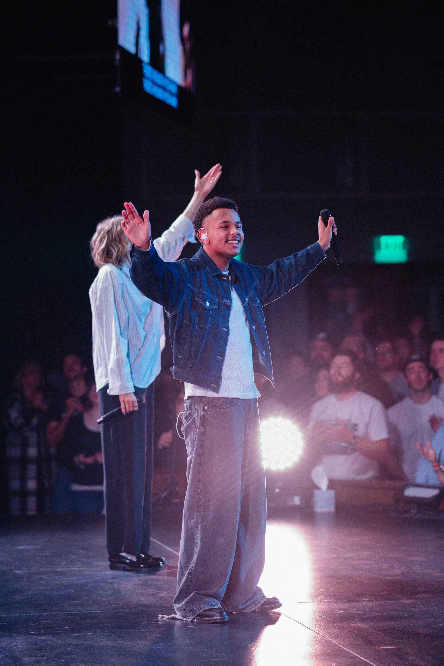 A young man on stage smiles with arms outstretched, holding a microphone.