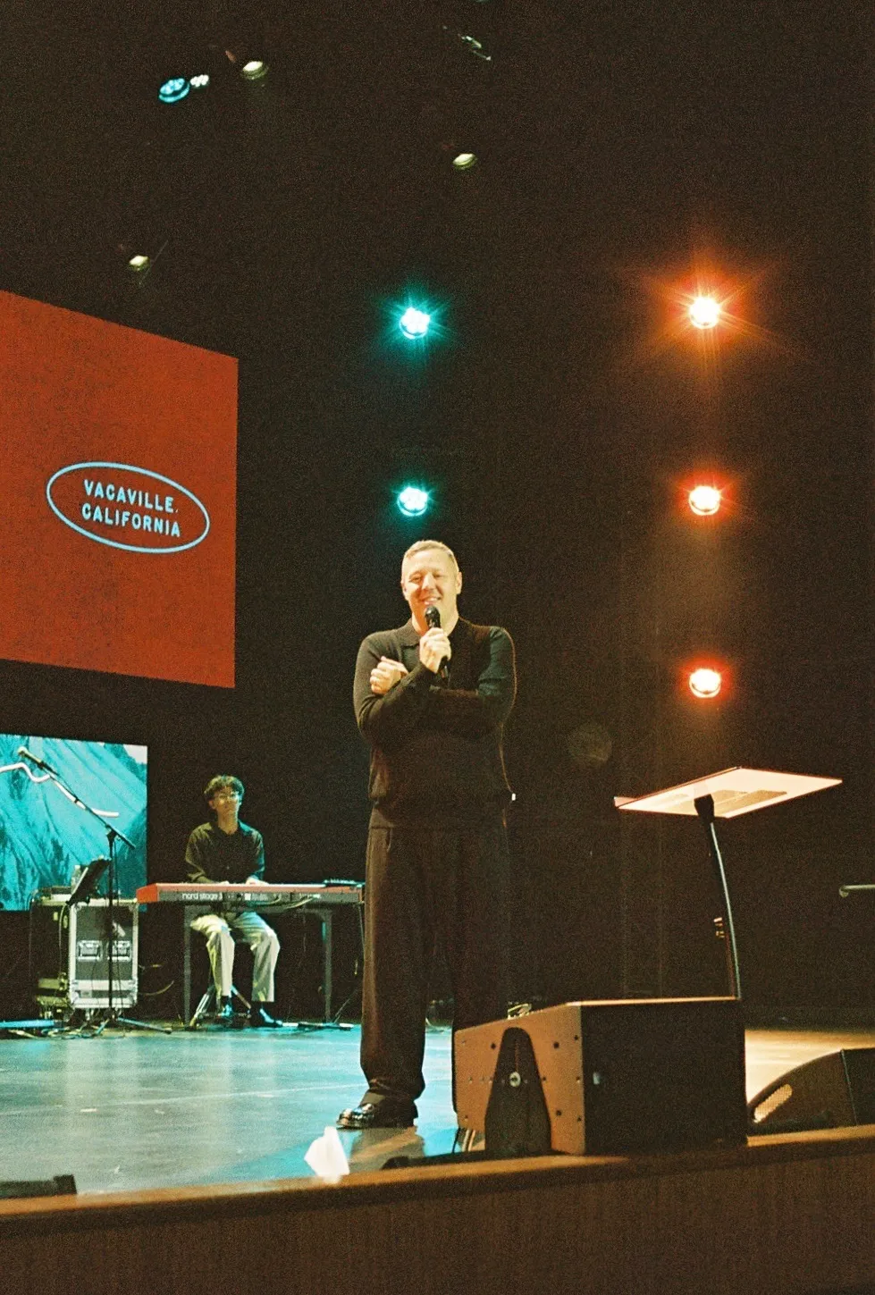 A man speaks on stage in front of a screen reading "Vacaville, California," with another musician seated at a keyboard behind him.