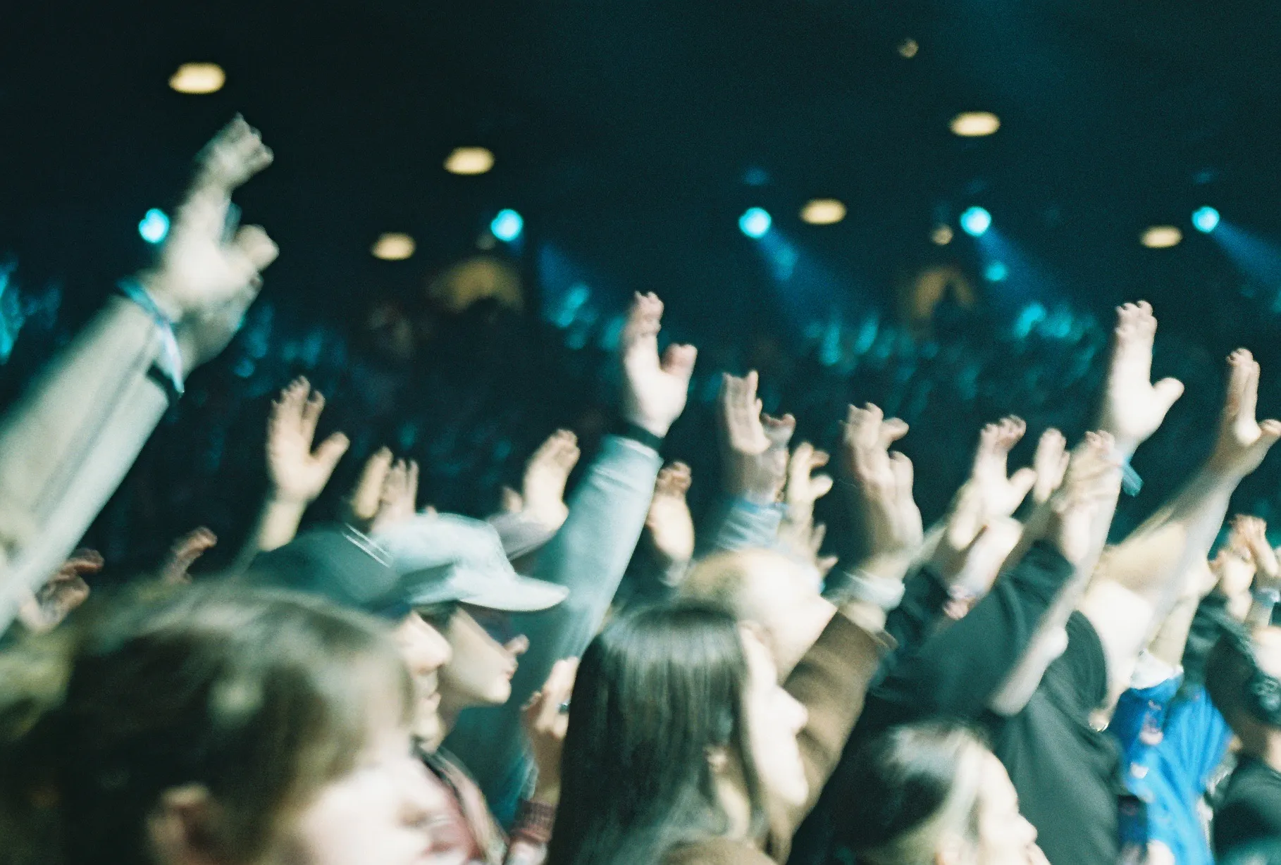 A blurred film photo of a crowd with hands raised, lit by teal stage light.