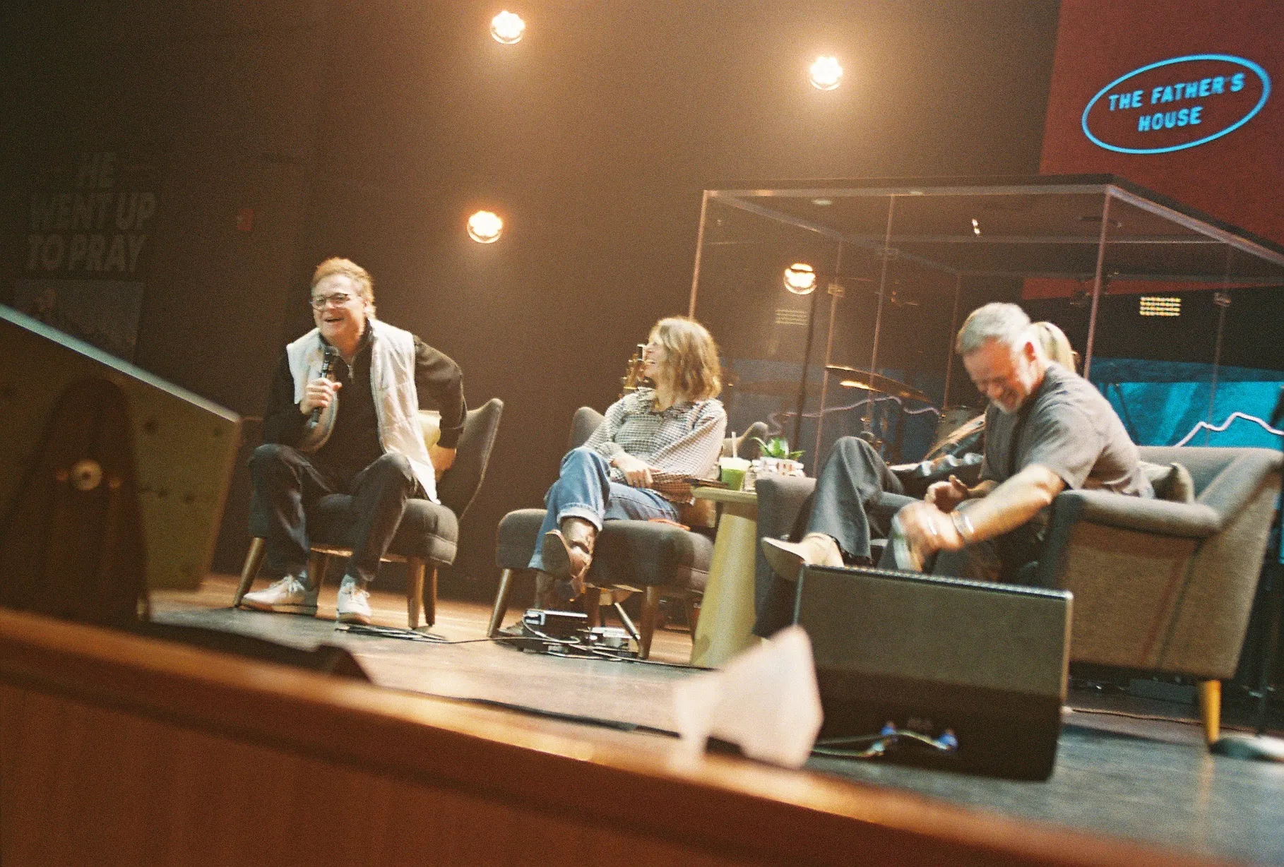 A film photo of three people seated in armchairs on stage, laughing together in conversation, with "The Father's House" sign glowing behind them.