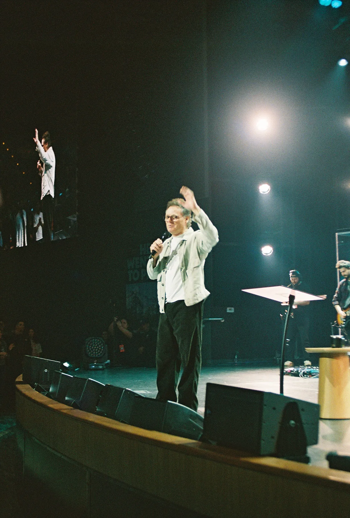 A film photo of a man on stage with one arm raised, speaking into a microphone, with another figure visible on a screen above.