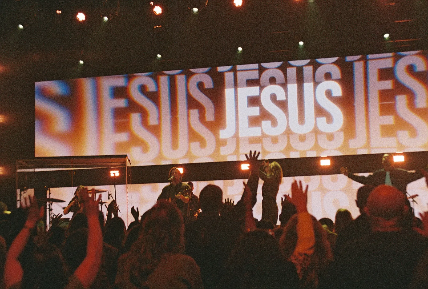 A film photo of a crowd with hands raised in front of a screen reading "JESUS" repeated in stylized type.