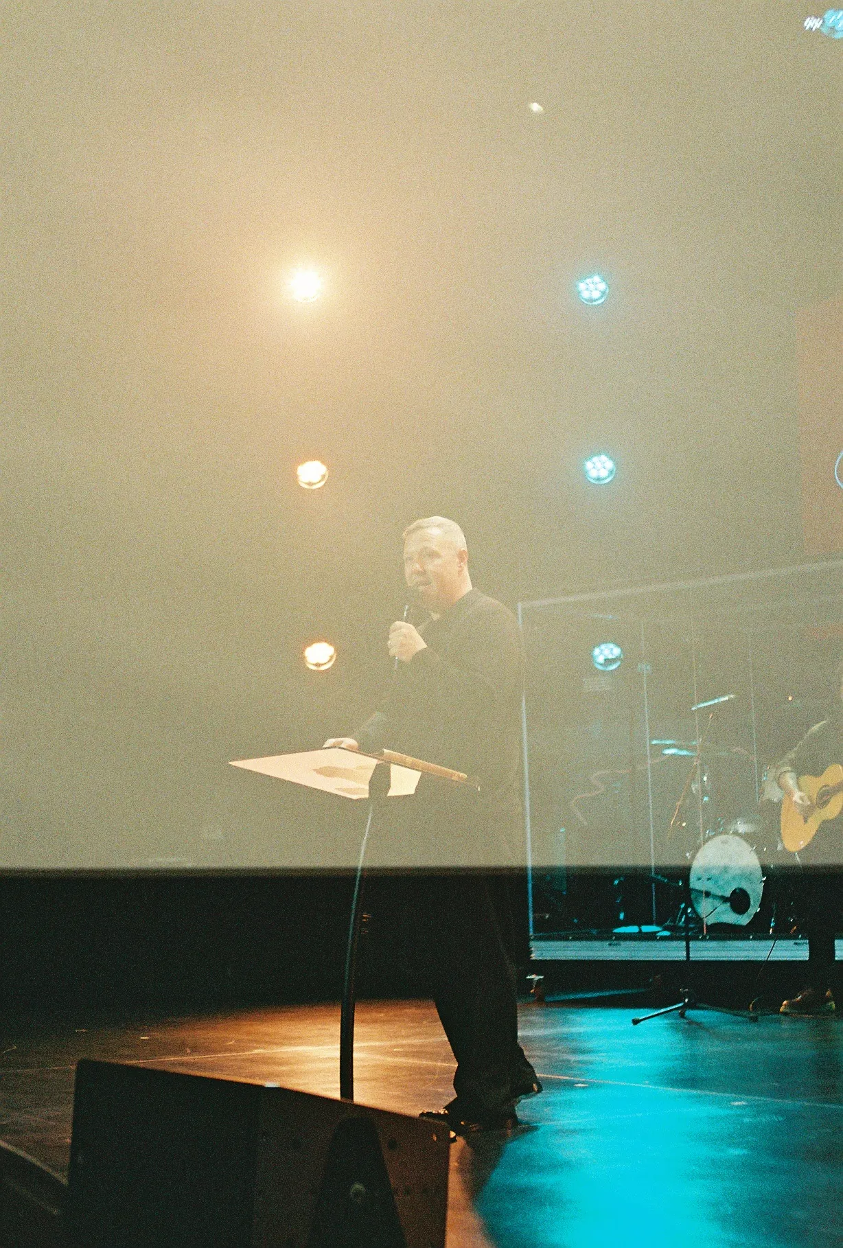 A film photo of a man speaking at a podium on a dim stage lit by colored stage lights.